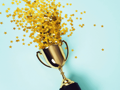 Golden award on a blue table with golden confetti stars spilling out of it.