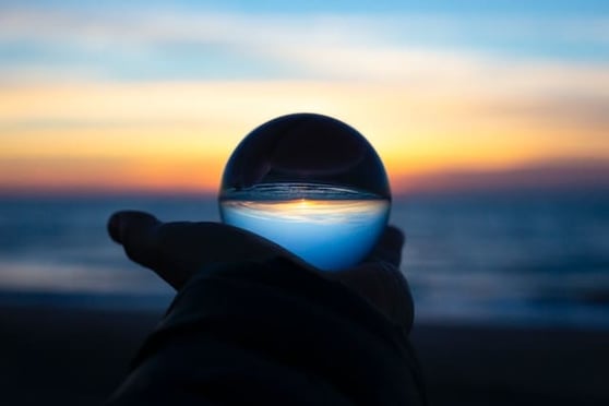 Hand holding a crystal ball in front of a beach at sunset.