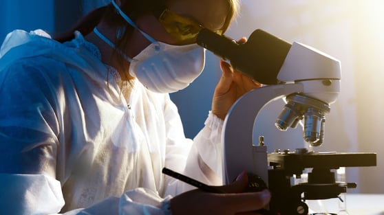Person wearing labcoat and mask looking into a microscope.