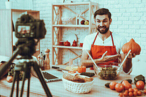 Man standing by a kitchen table filled with pots, talking into a camera standing on a tripod