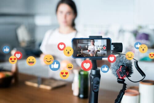 Woman in the background with a phone on a tripod in the foreground, with floating emojis.