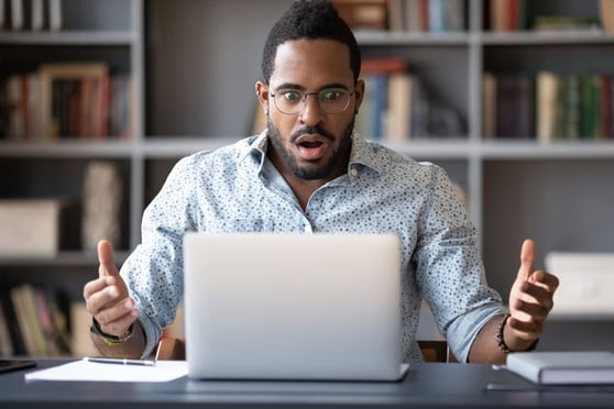 Man sitting at a desk in front of his laptop, arms spread and mouth open in surprise.