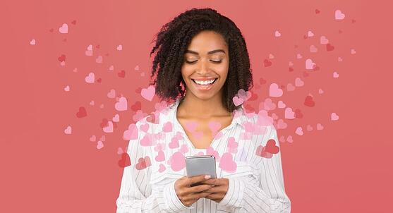 Smiling woman looking at her phone, with flying pink and red hearts coming out of her phone.