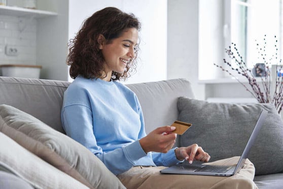 A smiling woman sitting on a couch, with a laptop on her lap and a credit card in her hand.