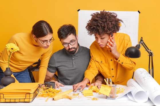 3 people crowded around a desk with notes, yellow background.