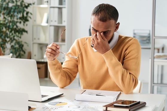 Man in yellow sweater sitting in front of reports, holding the bridge of his nose in frustration.