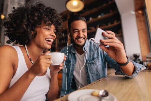 Two people sitting at a coffee bar, laughing at a cell phone.